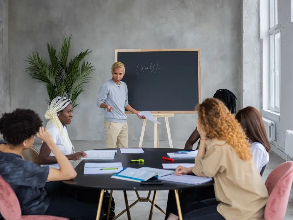 Students working together on math at a table with a teacher and chalkboard